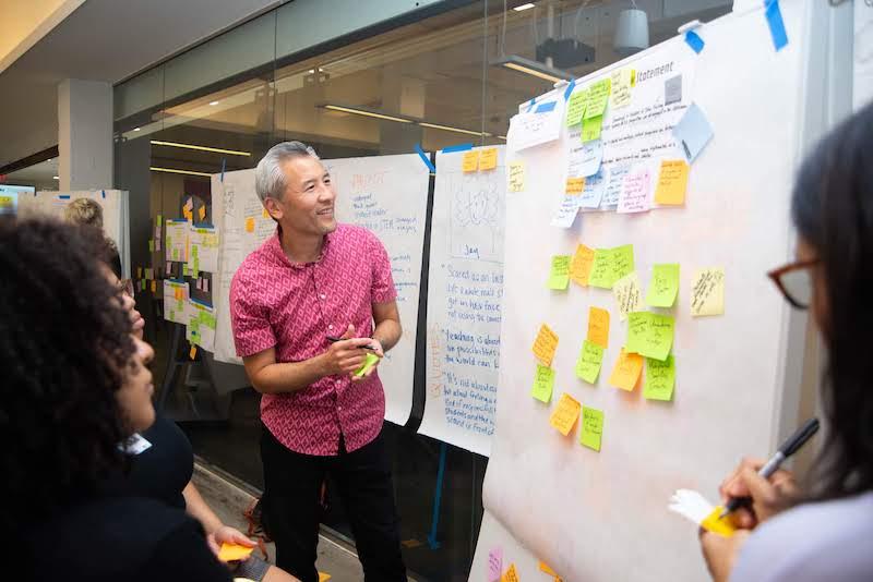 Person smiling holding post-its notes looking at a whiteboard with two other people